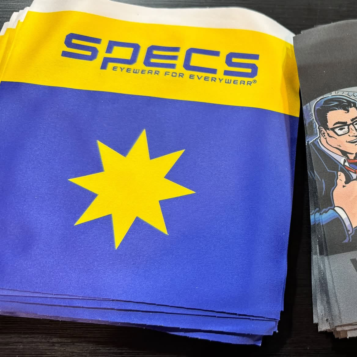 A stack of blue and yellow Specs eyewear packaging napkins on a dark wooden surface, with a printed card featuring a cartoon character partially visible on the right.
