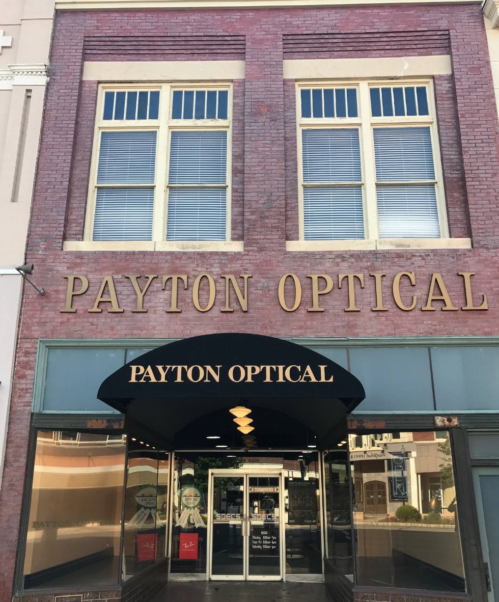 Front exterior of a brick building with large gold 'PAYTON OPTICAL' lettering above the entrance and a black awning with matching lettering.