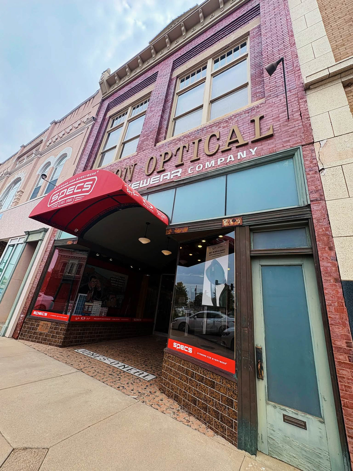 The storefront of Specs Rx Hutchinson featuring a red brick facade, a red awning with the Boebs logo, and a light blue-green wooden door on a city sidewalk.