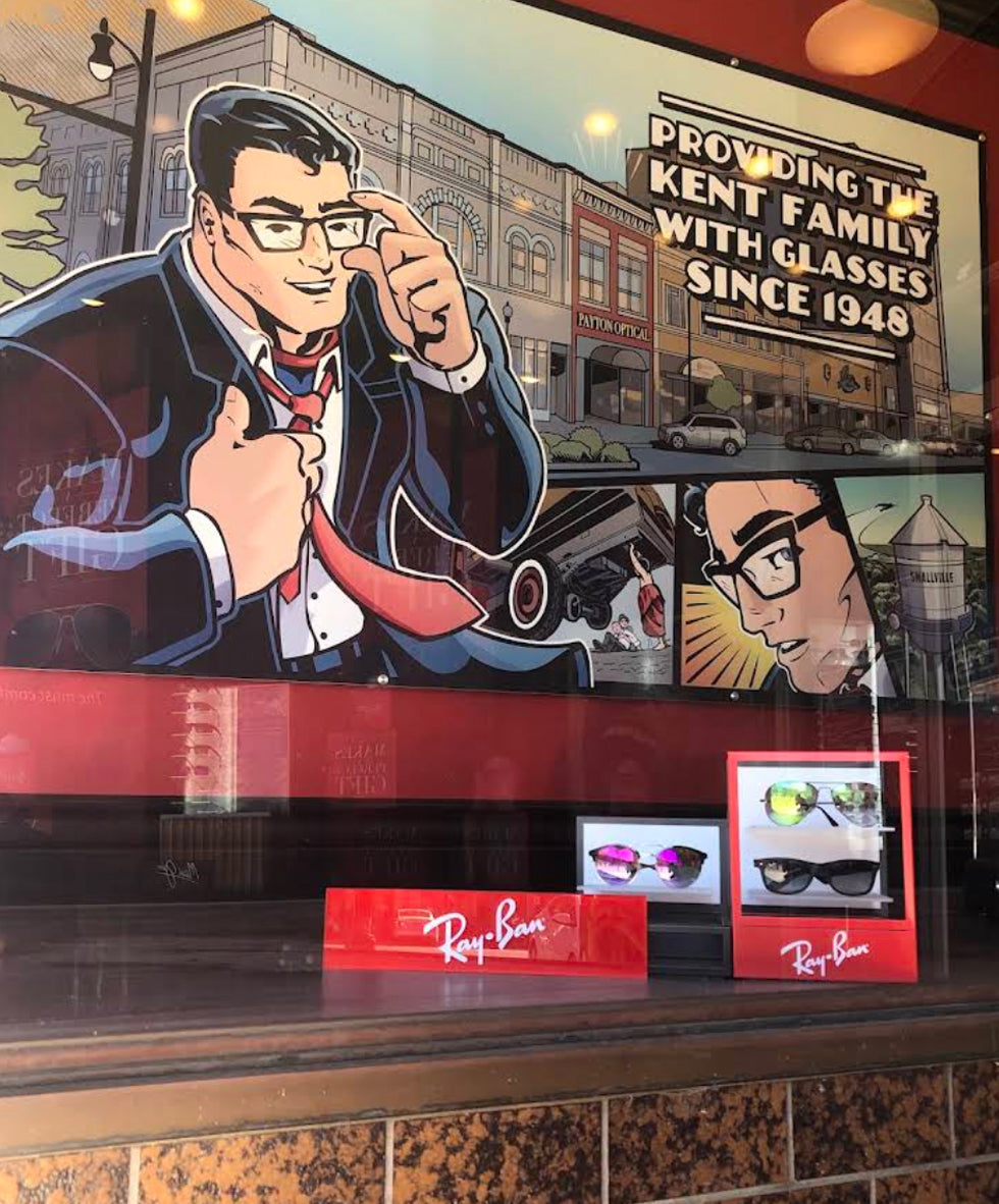 A Ray-Ban display inside the Specs Rx Hutchinson Kansas store window featuring a large vintage-style comic mural of superman in a suit and glasses, with two small Ray-Ban sunglasses boxes on the counter in front. Smallville Kansas