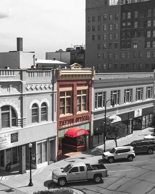 A selective-color photograph of a city street corner featuring a prominent red brick building with a red awning labeled 'PAYTON OPTICAL' which is now Specs Rx Hutchinson. The surrounding buildings, parked cars, and street are rendered in black and white, creating a strong contrast that highlights the red storefront.